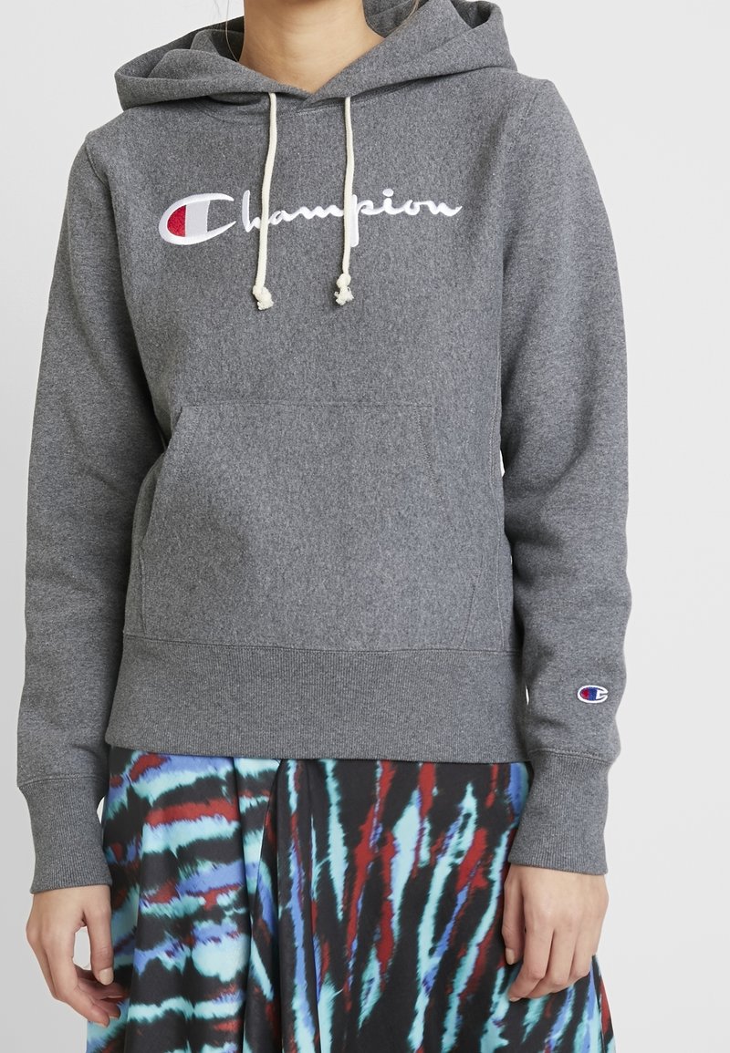Personne portant un sweat à capuche Champion gris avec des cordons blancs et une jupe tie-dye multicolore sur un fond uni.