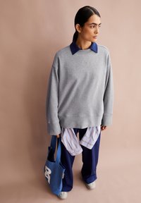 Person mit dunklen Haaren im Pferdeschwanz, trägt einen grauen oversized Sweatshirt, marineblaue Hose, weiße Sneakers, hält eine blaue Tragetasche und schaut nach rechts.