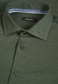 Camicia verde con texture, dotata di colletto, bottoni neri e polsini interni decorati con cerchi blu e bianchi. Etichetta del marchio visibile.