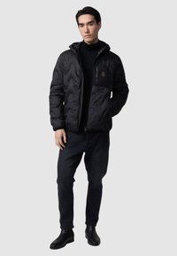 Giacca a soffietto nera con cappuccio, caratterizzata da un motivo cucito e una tasca con zip. Indossata sopra un dolcevita nero e pantaloni scuri.