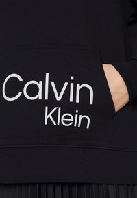 Ruka zastrčená v přední kapse černé mikiny Calvin Klein s bílým logem značky na látce.
