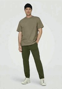 Olive green cotton t-shirt paired with dark green pants and white sneakers. The model wears a black knit beanie and stands upright.