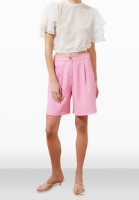 Blusa bianca con volant e pois abbinata a pantaloni corti rosa a pieghe. L'outfit presenta un design corto e una chiusura frontale con un solo bottone.