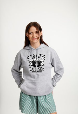 Ung kvinde med brunt hår iført en grå Star Wars-hættetrøje og grønne stribede shorts, stående med hænderne i lommerne mod en hvid baggrund.