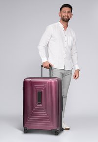 Hartschalenkoffer in einem tiefen Lila mit horizontalen Rillen, schwarzen Akzenten, einem ausziehbaren Griff und vier Rollen für Mobilität.