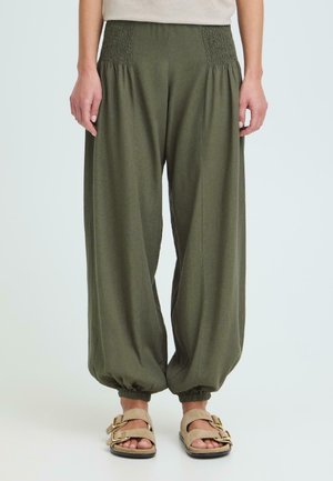 Løse olivengrønne bukser med elastisk midje og ankler, brukt med beige sandaler med dobbel stropp på lys bakgrunn.