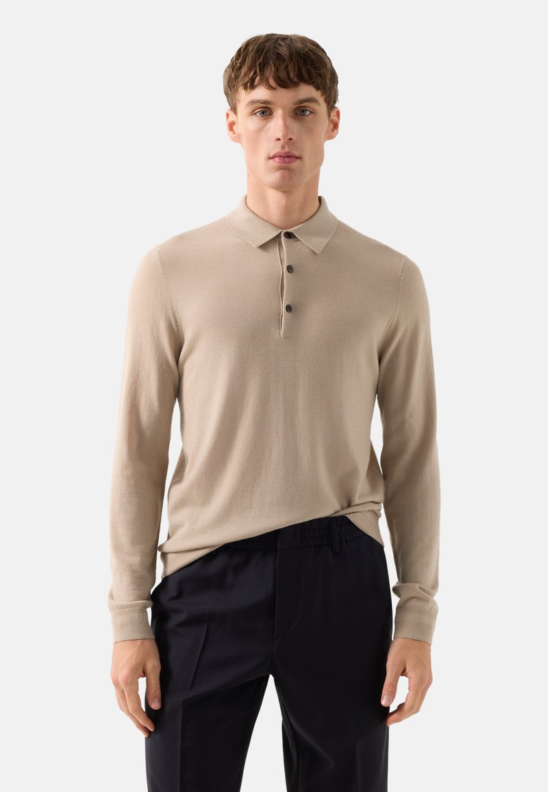 Jonge man die een beige poloshirt met lange mouwen en zwarte broek draagt, staand tegen een effen lichte achtergrond.