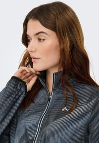 Giacca con cerniera a righe in tonalità scure, con collo alto, rifiniture nere e dettaglio di branding discreto sul petto. Materiale leggero e testurizzato.