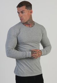 Camiseta de manga larga gris hecha de un tejido suave y elástico; cuenta con mangas ajustadas y un logo sutil en el pecho. El modelo posa con los brazos cruzados.