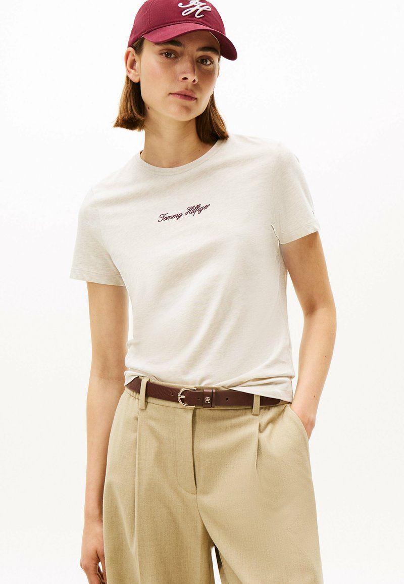 Hvid bomulds-T-shirt med "Tommy Hilfiger" broderet i mørkerød. Kombination med beige plisserede bukser og et brunt bælte. Mørkerød kasket.