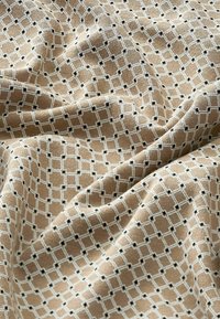 Tissu beige avec un motif répété de grille en losange blanc et de petits points noirs aux intersections, présenté avec des plis et une texture visible.