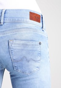 Vaqueros de mezclilla de color azul claro, con un parche de cuero con el logo, bolsillo trasero con costura en patrón de olas y detalles estándar en el cinturón.