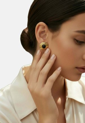 Gold-toned round earrings with spiral detailing and a central black stone, paired with a light satin blouse and a sleek hairstyle.