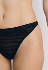 Crni čipkasti tanga gaćice s horizontalnim prugastim uzorkom i glatkim pojasom. Brand "Sloggi" suptilno je otisnut na pojasu.