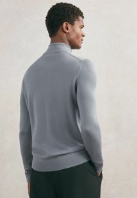 Mężczyzna stojący bokiem, ubrany w dopasowany jasnoszary sweter z golfem i czarne spodnie, z rękami w kieszeniach na jednolitym tle.