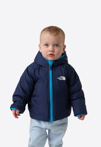 Vastsündinu siniste silmadega, kandes tumelilla ja türkiissinist kapuutsiga jopet ja helehalli pükse, seisab ühevärvilise tausta ees.