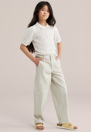 Fiatal lány áll zsebekkel a kezében, fehér rövid ujjú kötött pólót, bézs színű bő szárú nadrágot és sárga szandált visel, egyszerű háttér előtt.