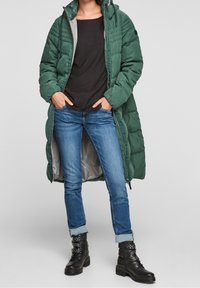 Długa zielona kurtka puchowa z kapturem, szarym podszyciem i przeszyciami na górze. W zestawie z ciemną koszulą, spodnie dżinsowe z podwiniętymi mankietami i czarnymi butami na sznurówki.