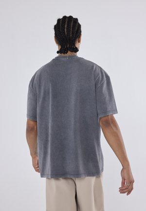 Person med flettet hår iført en løs grå T-shirt med korte ærmer og beige bukser, set bagfra mod en ensfarvet baggrund.