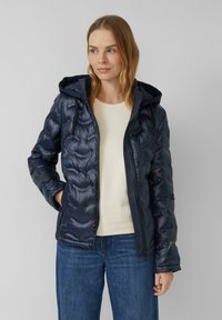 Doudoune marine avec capuche, présentant un design matelassé, deux poches latérales et une texture lisse. Portée par-dessus un haut de couleur claire.
