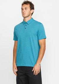 Tanzanienfarbendes Poloshirt aus strukturierter Baumwolle mit klassischem Kragen, drei schwarzen Knöpfen und kurzen Ärmeln. Kombiniert mit schwarzen Hosen.