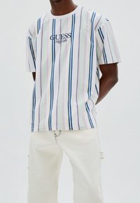 Camiseta de manga corta en blanco con rayas pastel en rosa, azul y verde. Presenta el logo de "GUESS Originals" en el pecho. Combínala con pantalones claros.