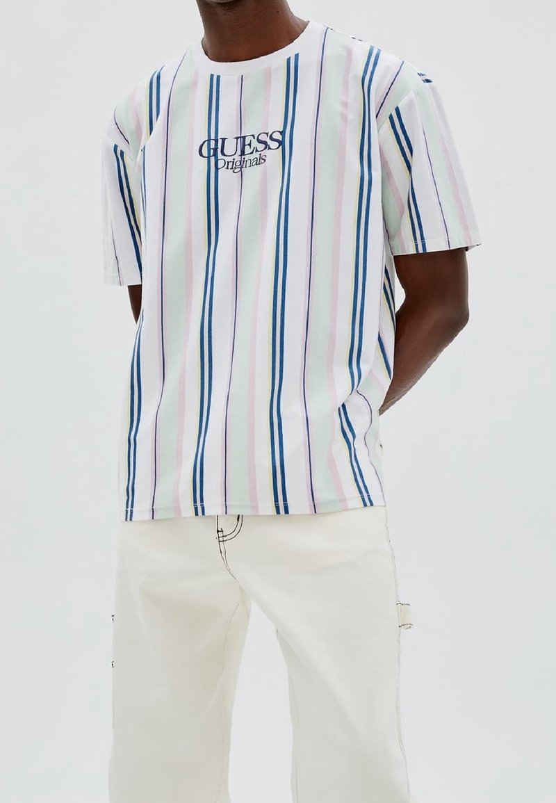 Camiseta de manga corta en blanco con rayas pastel en rosa, azul y verde. Presenta el logo de "GUESS Originals" en el pecho. Combínala con pantalones claros.