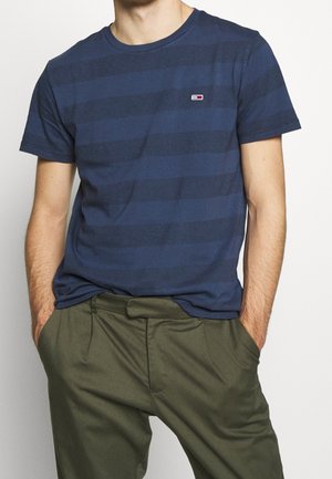 Mand iført marineblå, stribet t-shirt med small rektangulært logo, og olivengrønne bukser med hænderne i lommerne.