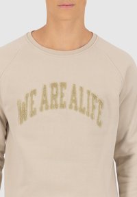 Beige sweatshirt met raglanmouwen en metallic gouden textuur lettering die "WE ARE ALIFE" op de borst zegt.