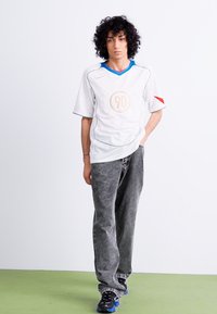 Camiseta deportiva blanca de manga corta con acentos azules y rojos, que presenta un diseño circular de "90" en el centro. Combinada con pantalones oscuros y texturizados.
