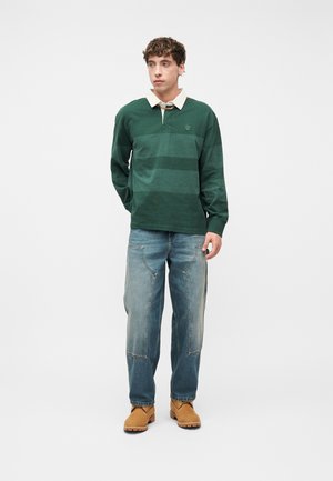 Ung mann stående iført grønnstripet langermet polo, løse blå jeans med frontlommer, og brune arbeidssko mot hvit bakgrunn.