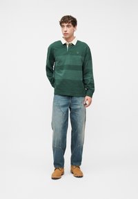 Νεαρός άνδρας που στέκεται, φοράει μακρυμάνικο πουλόβερ πόλο με πράσινες ρίγες, φαρδιά μπλε τζιν με μπροστινές τσέπες και ταμπά εργασιακές μπότες σε λευκό φόντο.