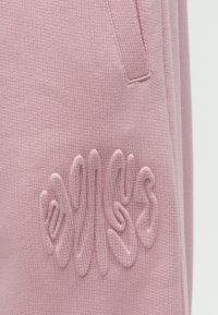 Calças de treino cor-de-rosa em tecido polar com um logótipo texturado e bordado na coxa esquerda. Apresentam um tecido macio e malhado, com bolsos laterais.