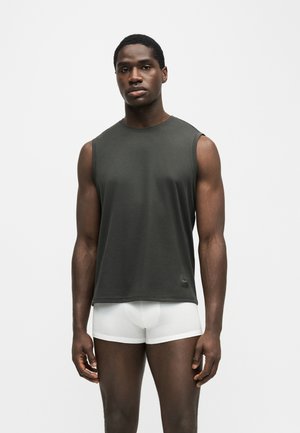 Homme à la peau foncée debout, portant un débardeur gris foncé et un boxer blanc, sur un fond blanc uni.