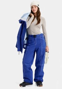 Γυναίκα με λευκό σκουφάκι, μπεζ μπλούζα μακρυμάνικη, μπλε παντελόνια χιονιού, μαύρες μπότες χιονιού, κρατώντας μπλε μπουφάν και λευκά γάντια.