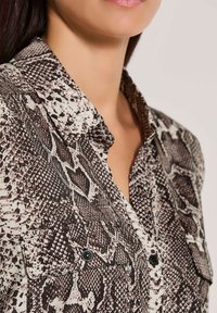 Braun-weiße Schlange Druckhemd mit Kragen, ausgestattet mit einer Brusttasche und schwarzen Knopfakzenten. Glatte Stoffoberfläche.