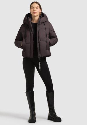 Mujer de pie con las manos en los bolsillos, vistiendo una chaqueta acolchada negra, leggings negros y botas negras hasta la rodilla sobre un fondo liso.