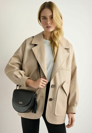 Blondynka w beżowej, oversize'owej kurtce, białym topie i czarnych spodniach, trzyma czarną skórzaną torebkę na ramię z frędzlami na białym tle.