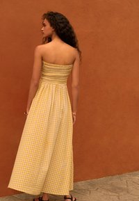 Donna con capelli ricci che indossa un lungo vestito senza spalline a quadretti gialli e bianchi, in piedi contro un muro arancione con texture.