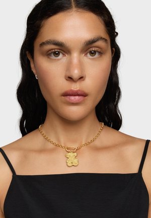 Collana in oro con pendente a forma di orsetto dal finitura texturizzata, indossata su un collo nudo, in contrasto con una maglietta nera.