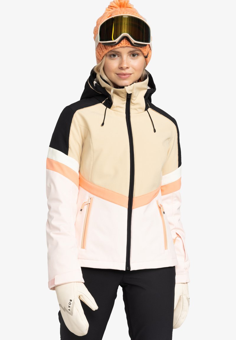 Ľahká lyžiarska bunda v béžovej, čiernej a ružovej farbe s vreckami na zips, nastaviteľnou kapucňou a dizajnom s farebnými blokmi. Nosí sa s bielymi rukavicami.