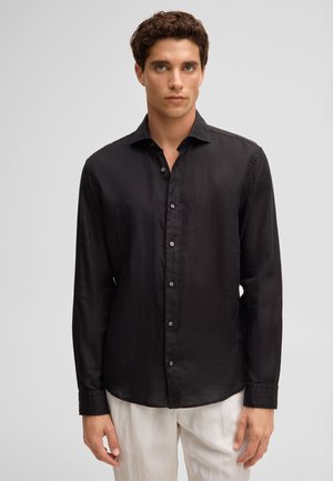 Jeune homme aux cheveux bouclés porte une chemise noire à manches longues boutonnée et un pantalon blanc, debout sur un fond clair uni.