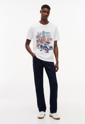 Noor mees valges Miami graafilise trükiga T-särgis, kus kujutatud võidusõiduauto ja palmipuud, tumedad teksapüksid ja valged tossud, seisab ühe käega taskus.