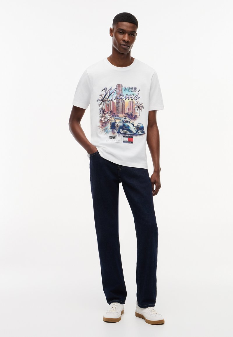 Ung mand iført hvid Miami T-shirt med grafik af en racerbils og palmer, mørke jeans og hvide sneakers, stående med den ene hånd i lommen.