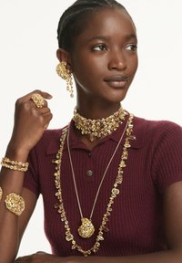 I gioielli in oro presentano un anello floreale spesso, collane a strati e grandi orecchini con vari gemme. Il modello indossa un polo bordeaux.