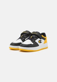 Sorte og hvite sneakers med et gult element. Har en glatt lærøverdel, rund tå, polstret krage og gummisåle med teksturert grep.