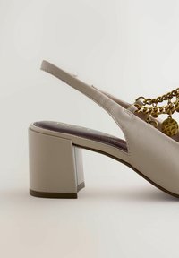 Chaussure à talon bloc beige à bride arrière avec détail de chaîne dorée et breloque sur fond neutre, vue de profil latéral et talon.