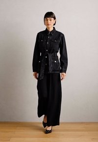 Svart denimjakke med belte i midjen, frontknapper og to brystlommer, sammen med et langt svart skjørt og prikkete svarte sko.