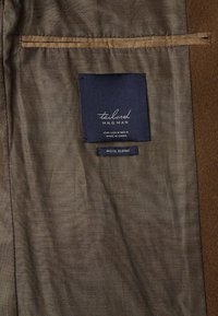 Indumento in misto lana con una superficie marrone e testurizzata. Presenta un'etichetta scura con "tailored MNG MAN" e istruzioni per la cura, vicino a una tasca.