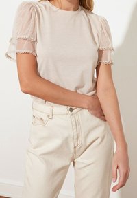 Top beige chiaro con maniche corte a sbuffo trasparenti e rifiniture in pizzo, abbinato a jeans in denim chiaro a vita alta con cuciture arancioni.